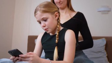 Sevecen anne kızının saçlarını örerken genç kız akıllı telefonuyla meşgul oluyor sıcak ve huzurlu bir yatak odasında rahat bir şekilde yatıyor.