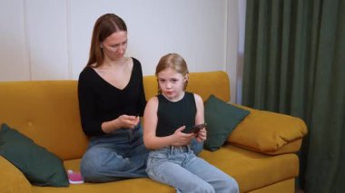 Anne sarışın kızın saçını dikkatlice örerken çocuk tabletin tadını çıkarıyor, rahat oturma odasındaki parlak sarı kanepede dinleniyor.