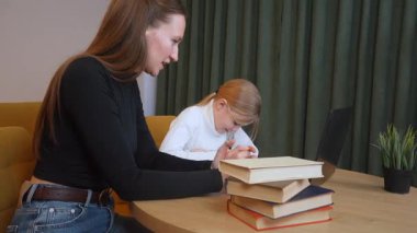 İlköğretim öğrencisi özenle not defterine yazarken, öğretmen kişisel rehberlik, dizüstü bilgisayar ve temel öğrenme desteği için referans kitapları sunuyor.