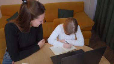 İlköğretim öğrencisi özenle not defterine yazarken, öğretmen kişisel rehberlik, dizüstü bilgisayar ve temel öğrenme desteği için referans kitapları sunuyor.