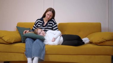 Annesinin, rahat sarı koltukta dinlenirken, kızının saçlarını hafifçe okşaması sessiz aile anlarında anne bakımını ve duygusal bağlantıyı somutlaştırması.