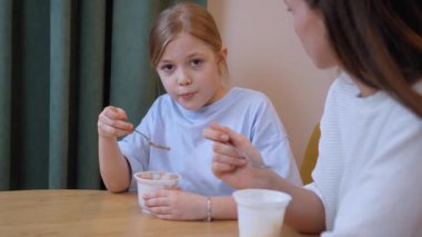 Sevecen anne, sabah kahvaltısı sırasında küçük kızı plastik bardaktan yoğurtla besliyor, ev mutfağında beslenme anını paylaşıyor.