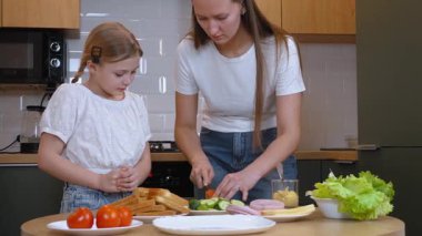 Annem kızıyla sandviç yaparken domatesi düzgünce dilimliyor, yemek yapma becerilerini gösteriyor ve modern mutfaklarında kaliteli aile zamanlarının tadını çıkarıyor.