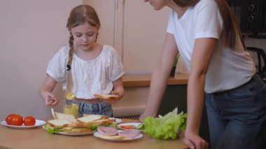 Sevgi dolu anne ve kız, ev yapımı sandviçler hazırlıyor, kremalı avokado yayıyor, taze marul, jambon ve peynir katmanlıyor, ortak bir neşe ve aşçılık bağıyla.
