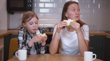 Mutlu aile, anne ve kız, mutfakta masada oturuyor ve kahvaltıda jambon, peynir, domates, salatalık ve marul ile sandviç yiyorlar.