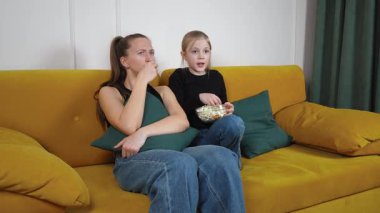 Yeşil yastıklı rahat sarı bir koltukta oturan anne ve genç kız, televizyonda korku filmi izliyor, patlamış mısır yiyor ve korkmuş görünüyorlar.