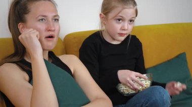 Yeşil yastıklı rahat sarı bir koltukta oturan anne ve genç kız, televizyonda korku filmi izliyor, patlamış mısır yiyor ve korkmuş görünüyorlar.