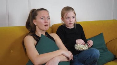Yeşil yastıklı rahat sarı bir koltukta oturan anne ve genç kız, televizyonda korku filmi izliyor, patlamış mısır yiyor ve korkmuş görünüyorlar.