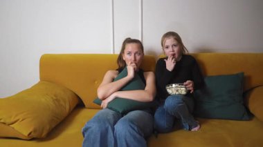 Anne ve kızı korku filmi izlerken gergin anlar yaşıyorlar, yastıklara sıkıca sarılıyorlar ve sarı oturma odasındaki kanepede cam kaseden patlamış mısır paylaşıyorlar.