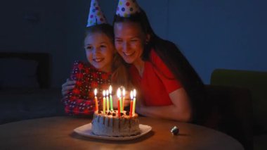 Sıcak mum ışığı, neşeli parti şapkaları takan genç bir kızı ve annesini aydınlatıyor, bir doğum günü pastasının etrafında toplanarak, aile kutlamalarının iç açıcı bir sahnesini yaratıyor.