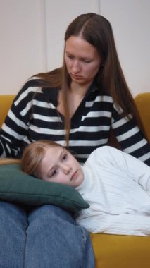 Anne, hasta kızını nazikçe rahatlatıyor, saçlarını okşuyor. Neşeli bir şekilde yüzünü okşuyor, moralini yükseltiyor ve rahat sarı kanepenin dikey videosunda duygusal destek sağlıyor.