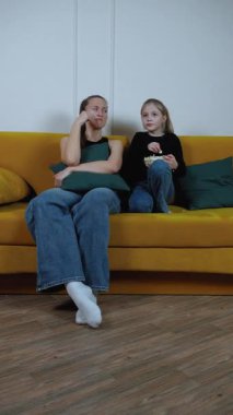 Anne ve kızı sarı kanepeye yakın oturuyor, yoğun film izlerken samimi bir anı paylaşıyor, çeşitli duygusal tepkiler sergiliyor ve patlamış mısırı birlikte dikey görüntülüyorlar.