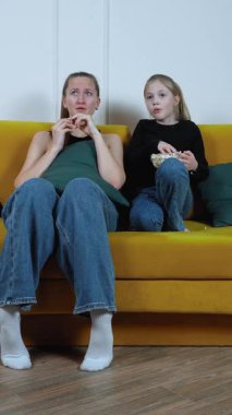 Anne ve kızı sarı kanepeye yakın oturuyor, yoğun film izlerken samimi bir anı paylaşıyor, çeşitli duygusal tepkiler sergiliyor ve patlamış mısırı birlikte dikey görüntülüyorlar.