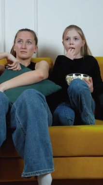 Anne ve kızı sarı kanepeye yakın oturuyor, yoğun film izlerken samimi bir anı paylaşıyor, çeşitli duygusal tepkiler sergiliyor ve patlamış mısırı birlikte dikey görüntülüyorlar.