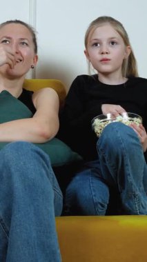 Anne ve kızı, rahat bir koltukta oturup, cam bir kaseden patlamış mısır yerken ve korkmuş yüz ifadelerini dikey olarak gösterirken televizyonda korku filmi izliyorlar.