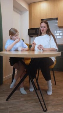 Anne ve kız yuvarlak bir masada modern bir mutfakta oturmuş, birlikte yemek yiyor, sağlıklı yoğurt yiyor ve canlı sohbetler yapıyor, aile bağlarının dokunaklı bir sahnesini yaratıyor.