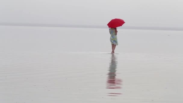 赤い傘のドレスを歩くビーチでエレガントな女性 海の反射と美しい女性 ビーチで美しい女性は 夏休みを楽しんで — ストック動画