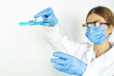 Koruyucu gözlüklü, maskeli ve eldivenli genç, çekici kadın bilim adamı bilimsel kimya laboratuarındaki bir pipetle test tüpüne mavi sıvı bir madde bırakıyor.   