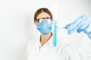 Koruyucu gözlüklü, maskeli ve eldivenli genç, çekici kadın bilim adamı bilimsel kimya laboratuarındaki bir pipetle test tüpüne mavi sıvı bir madde bırakıyor.   