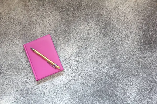 Pembe günlüğü bir gri beton zemin ve altın zarif tükenmez kalem. Üstten görünüm.