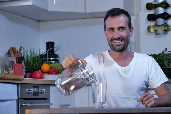 happy smiling man with stubble sitting at kitchen table and pouring water from jar into glass and looking at camera