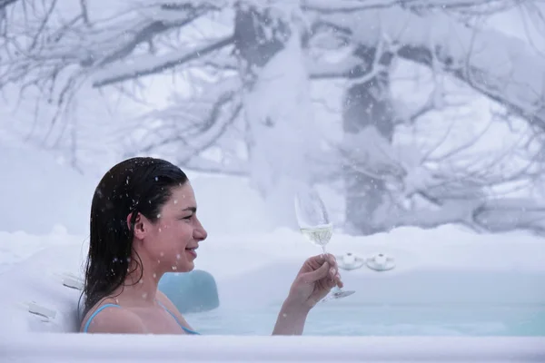 A beautiful woman drinks champagne in a hot tub surrounded by nature as the snow falls from the sky. Concept of: relaxation, nature, wellness and vacation.