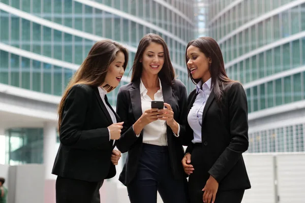 Three business women of different ethnicities (Asian, European and African American) look at a phone and smile. Concept of: social network, team, business people and female career.