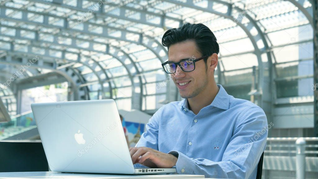 Young handsome businessman (student) in a blue shirt, at the table working with a laptop, serious, smiling. Concept: new business, business relationships, career growth, success, work online, station.