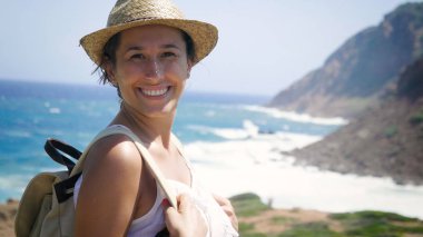 Portre, gülümseyen bir hasır şapkalı bir sırt çantası ile kaya, deniz ve dağlar arka yürüyüş bir güzel genç turist (kız), beyaz bir elbise içinde. Konsept: eğlence, güzel manzara.