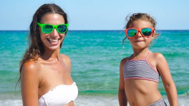 Anne ve küçük kızı eğleniyor, denizde, güneş gözlüğü, takıyor içinde mayolar, deniz mavi su arka plan gülümseyen iskambil.