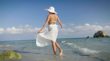 Güzel bir genç kız bir mayo bir hasır şapka, beyaz fuları, altın kum, deniz kıyısında bir arka plan dağlar ve deniz elini kolunu sallaya dönüyor. Konsept: deniz dinlenme, temiz hava, özgürlük, Güneş.