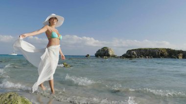 Güzel bir genç kız bir mayo bir hasır şapka, beyaz fuları, altın kum, deniz kıyısında bir arka plan dağlar ve deniz elini kolunu sallaya dönüyor. Konsept: deniz dinlenme, temiz hava, özgürlük, Güneş.
