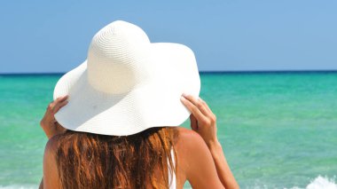 Yanık tenli bir kadın için deniz, mavi deniz suyu olan bir arka planda beyaz şapkalı, beyaz bir mayo içinde denize bakarak ile onu geri duruyor.