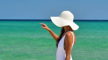 Yanık tenli bir kadın için deniz, mavi deniz suyu olan bir arka planda beyaz şapkalı, beyaz bir mayo içinde denize bakarak ile onu geri duruyor.