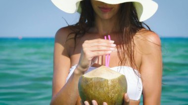 Güzel genç kız beyaz bir bikini, güneş gözlüğü, altın kum arka plan denizin kenarında bir Hindistan cevizi kokteyl içer. Konsept: deniz tatil, özgürlük, Güneş, seyahat, dalgalar, taze meyve, tatil, diyet, sıcak.