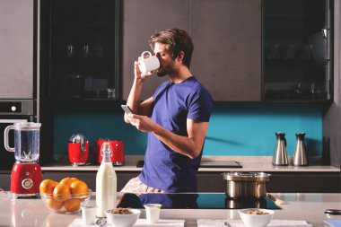 Bir adam Kupası'nda Kahvaltı ve yudumlarken çay veya süt görürken mutfakta bir ileti gönderir ya da ile belgili tanımlık telefon ve gülümsüyor çağırır. Kavramı: sosyal ağ, mesaj, teknoloji.