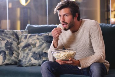 Bir adam televizyon önünde patlamış mısır yemek yerken kanepede oturan spor oyunu izler. Kavramı: spor, dinlenme, tantana, bahis.