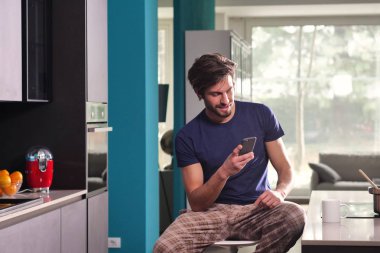 Bir adam Kupası'nda Kahvaltı ve yudumlarken çay veya süt görürken mutfakta bir ileti gönderir ya da ile belgili tanımlık telefon ve gülümsüyor çağırır. Kavramı: sosyal ağ, mesaj, teknoloji.