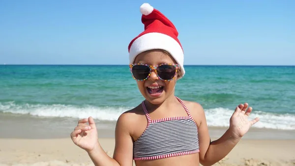 一个小女孩穿着圣诞老人的帽子 穿着游泳衣 戴着太阳镜 戴着海水蓝色的背景 在海上玩耍 — 图库照片
