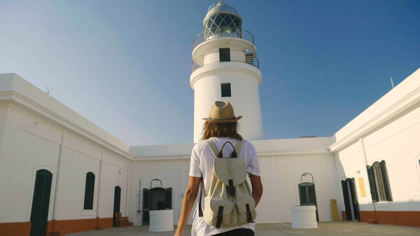 Portrait of a beautiful young tourist (girl), looks at an lighthouse, with a backpack, in a straw hat, a white house backdrop. Concept: recreation, beautiful view, sports, travel.