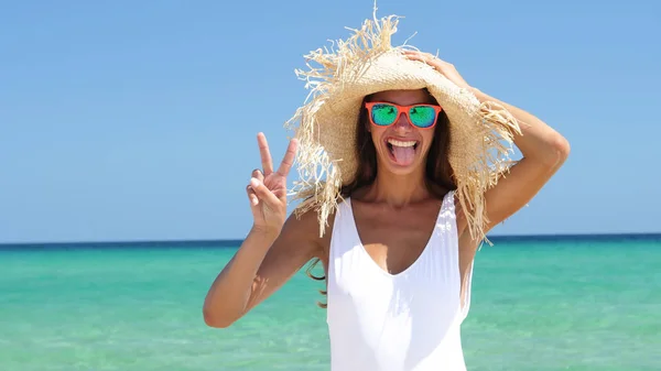 Beautiful happy woman at the sea, smiling in sunglasses, wearing a straw hat, in a white bathing suit, background of sea blue water.
