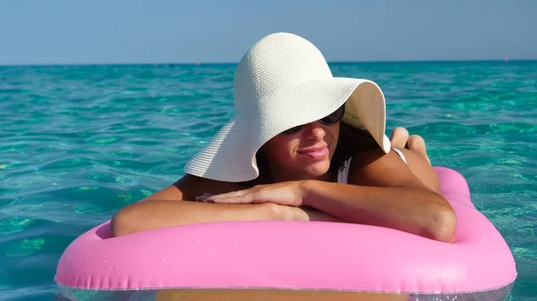 Beautiful woman, lady lies on a pink mattress in the water, smiling in a white hat, in a swimsuit and wearing sunglasses, a background of sea blue water.