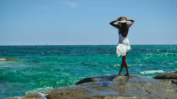 Kobieta Stoi Skałach Nad Morzem Słomkowym Kapeluszu Białej Sukni Noszenie — Zdjęcie stockowe
