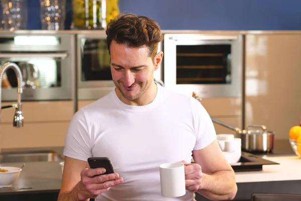 A man in the kitchen while having breakfast and sipping tea or milk in the cup sends a message or calls with the phone and smiles. Concept of: social network, message, technology.
