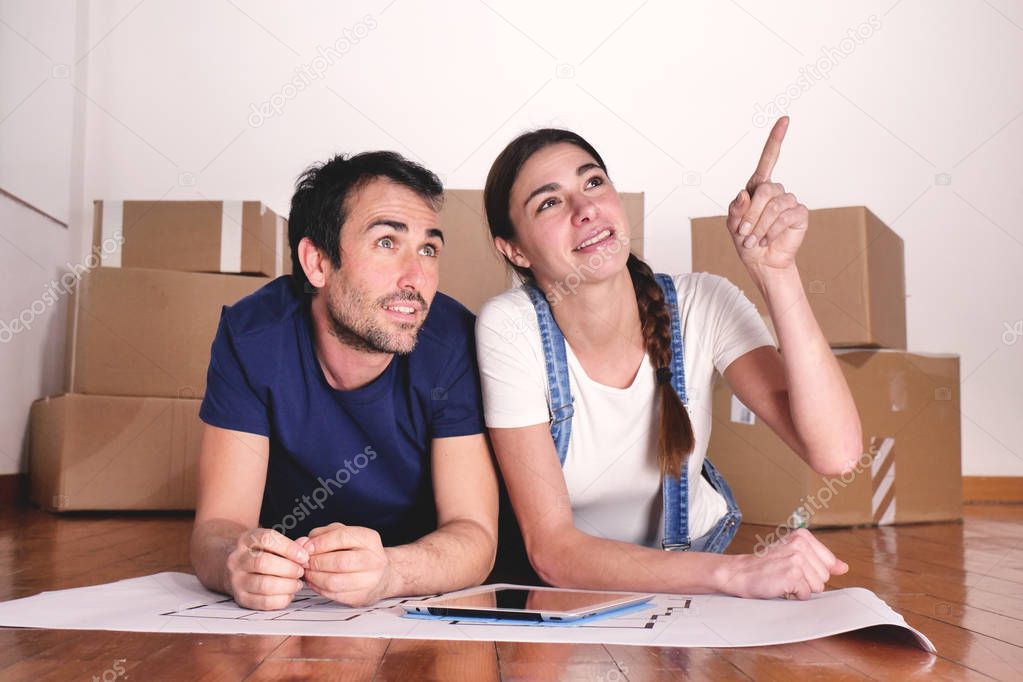 couple in love lying on wooden floor with room plan and digital tablet in new apartment, woman pointing finger 