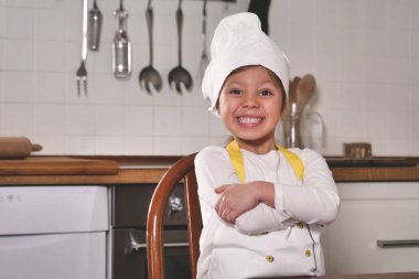 Küçük bir kızın portresi mutfakta profesyonel bir aşçı gibi giyinmiş unla oynuyor, ellerini kirletiyor ve mutlu gülümsüyor. Beslenme, aşçılık okulu, eğitim ve oyun kavramı.