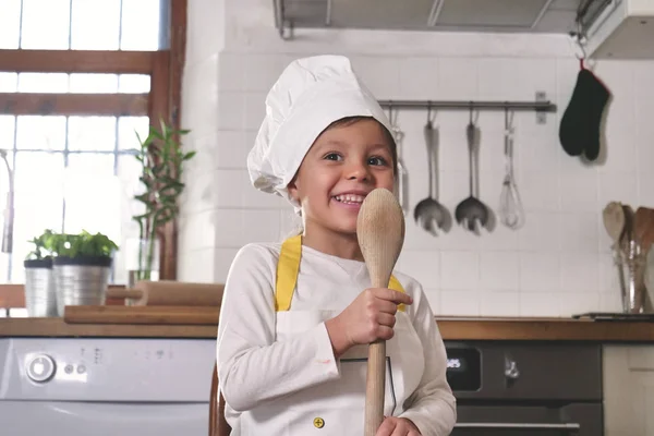 厨房里一个小女孩的肖像 她打扮成一个专业的厨师 面带微笑 手里拿着一个传统的木制厨房勺子 烹饪学校 教育和家庭 — 图库照片
