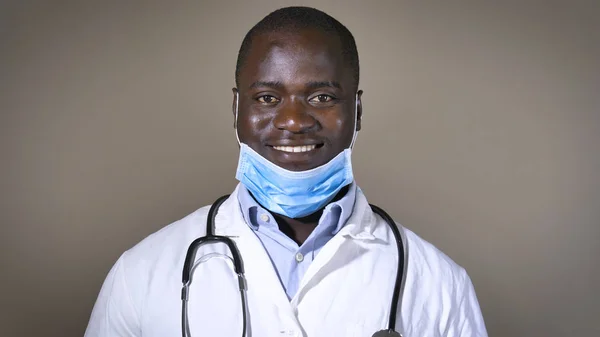 A successful doctor smiles (African American), in a respirator, in a white coat, stethoscope, brown eyes, brown background. Concept: science, beginning doctor, black skin, assistant, medic, profession