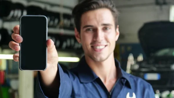 In a car garage a mechanic after checking the car smiles showing the phone for getting in touch using the app. Concept of: security, safety, insurance and professionalism, technology and applications.