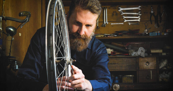 Well-groomed handsome bearded master hipster, specialist in bicycles, repairing a bicycle in his workshop, wheels, frame, spokes, the background of tools. Concept: pro bike, cycle passion, lifestyle.